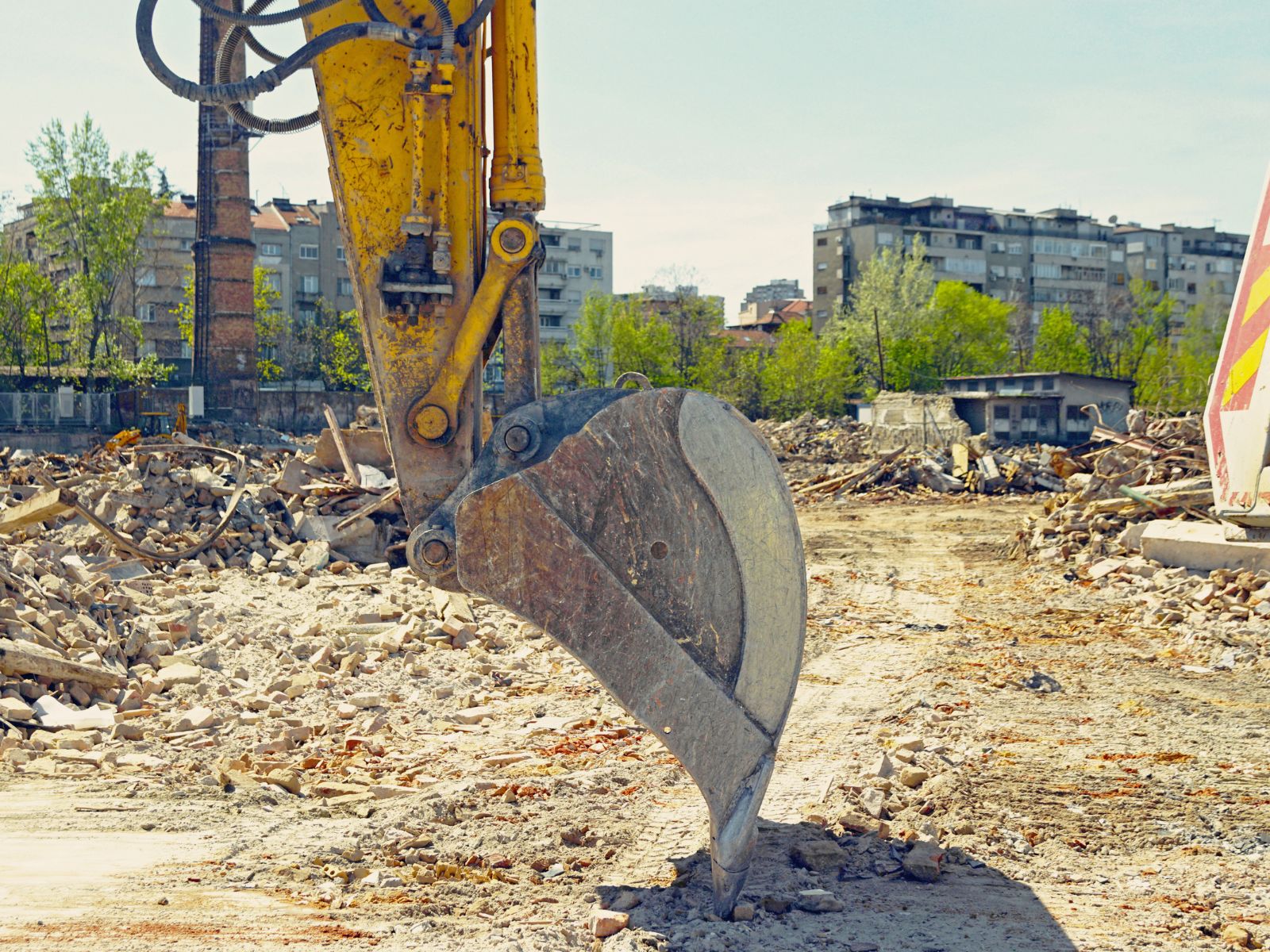 Close-up of a yellow excavator bucket at a demolition site with rubble and buildings in the background.