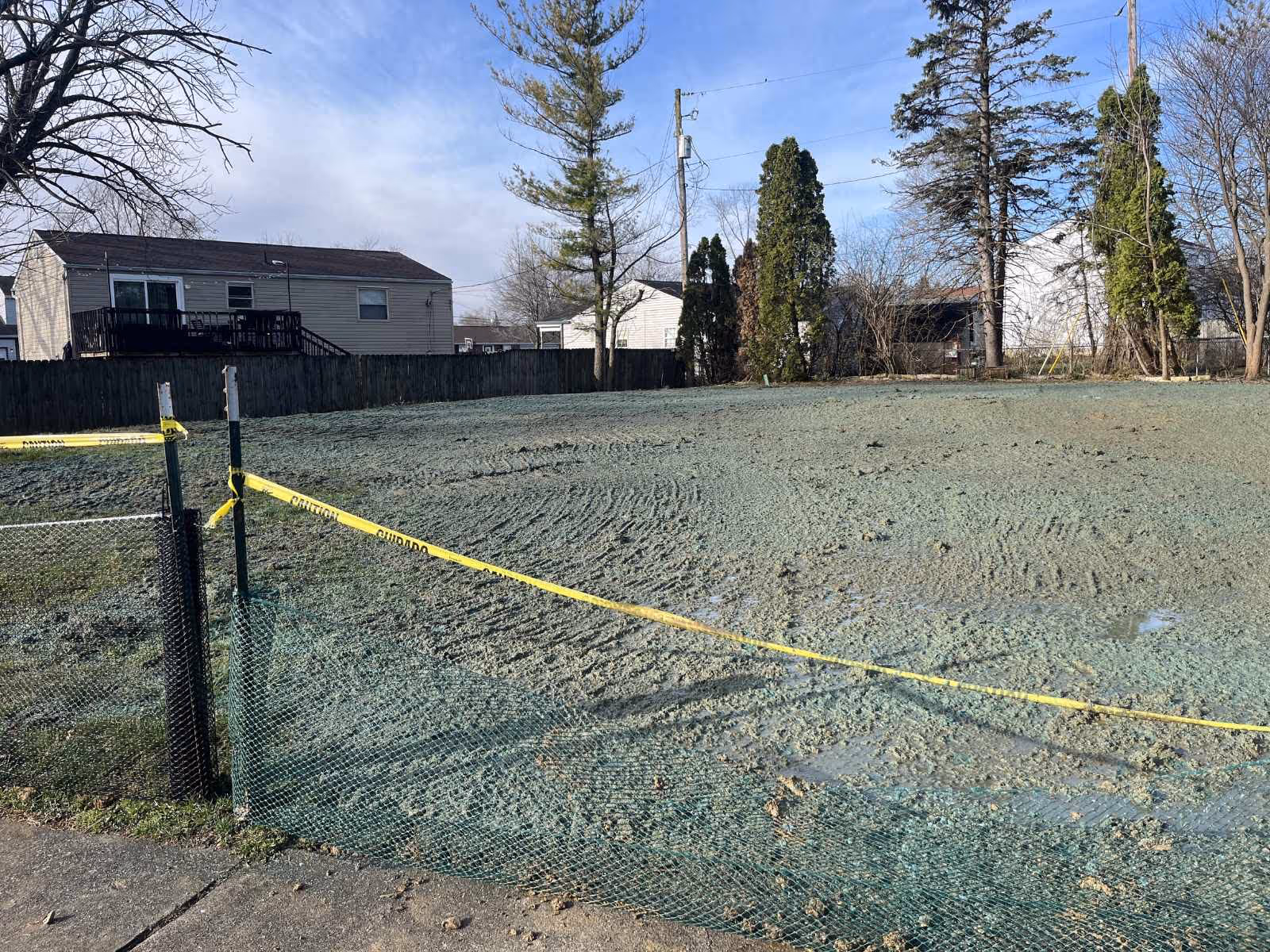 Open plot of land with freshly applied green hydroseeding material, surrounded by yellow caution tape and green mesh fencing.