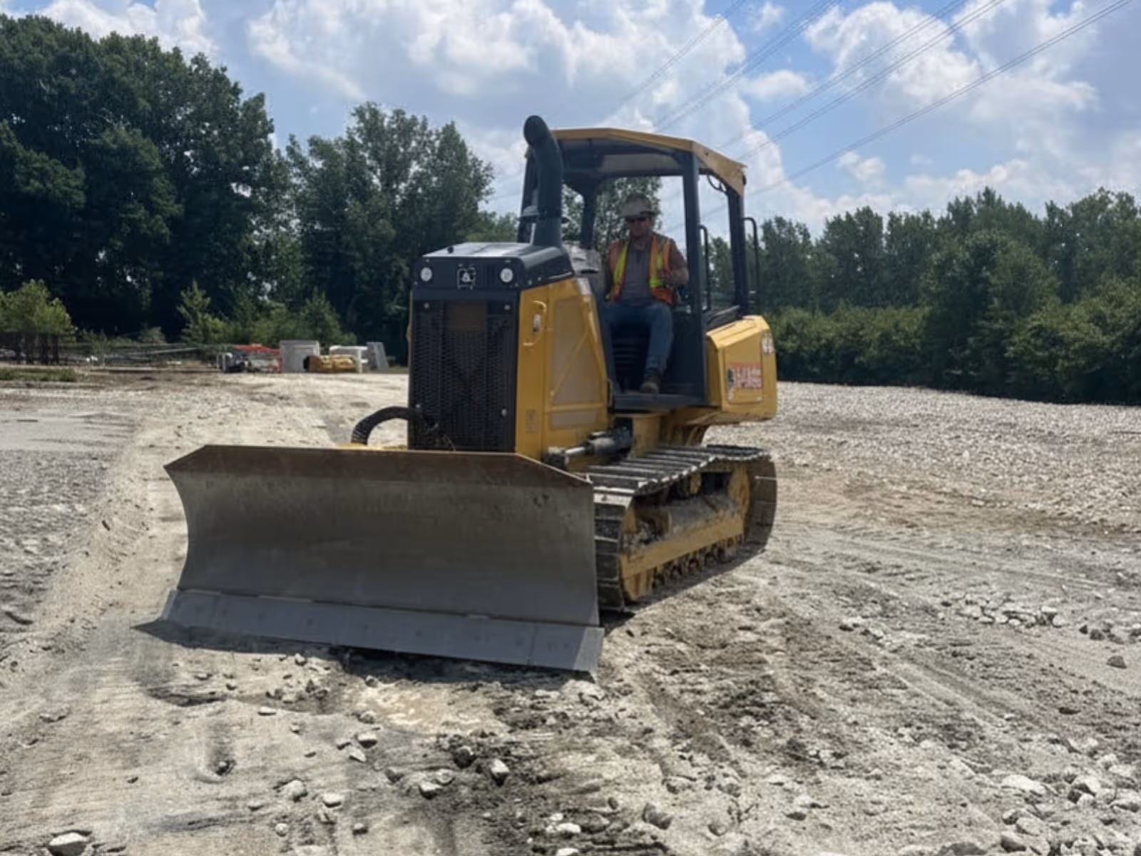 Construction worker operating a yellow bulldozer on a gravel and dirt surface with trees in the background.