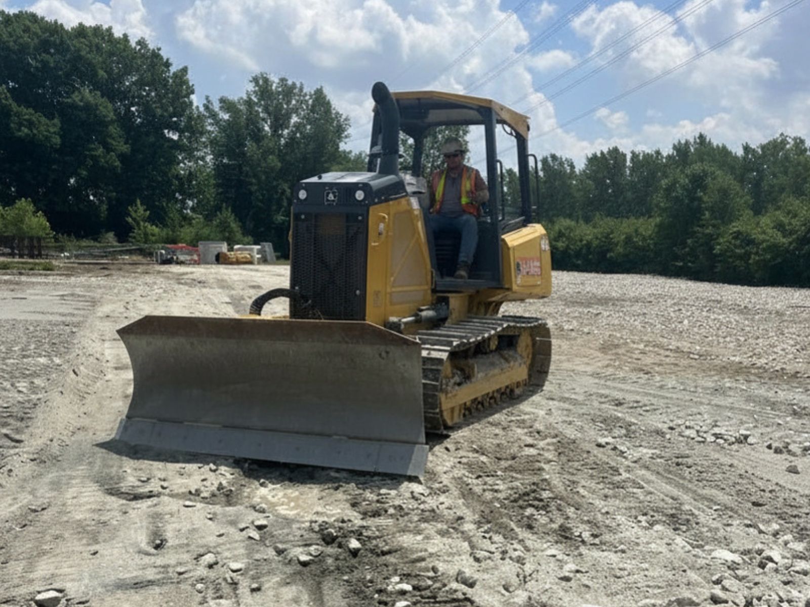 Construction worker operating a yellow bulldozer on a gravel and dirt surface with trees in the background.