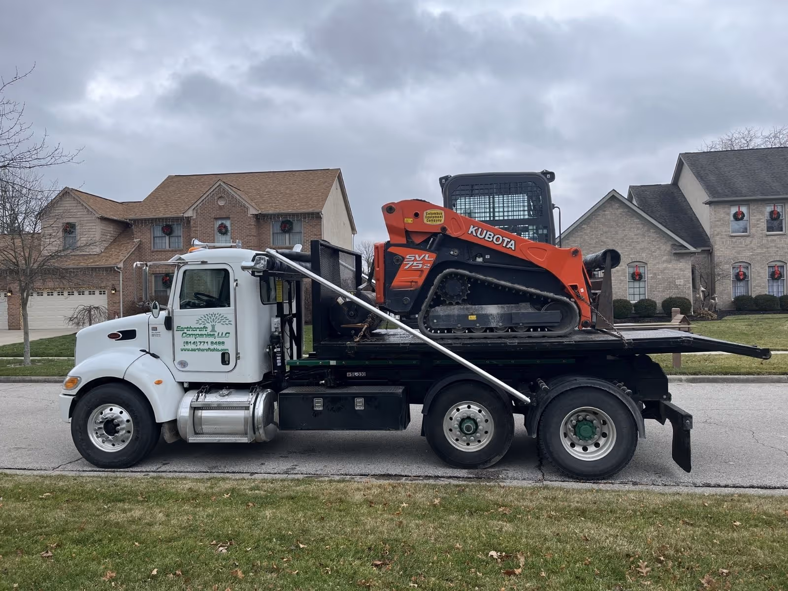 White flatbed truck from Earthworx Companies LLC carrying an orange Kubota SVL75-2 compact track loader parked on suburban street.