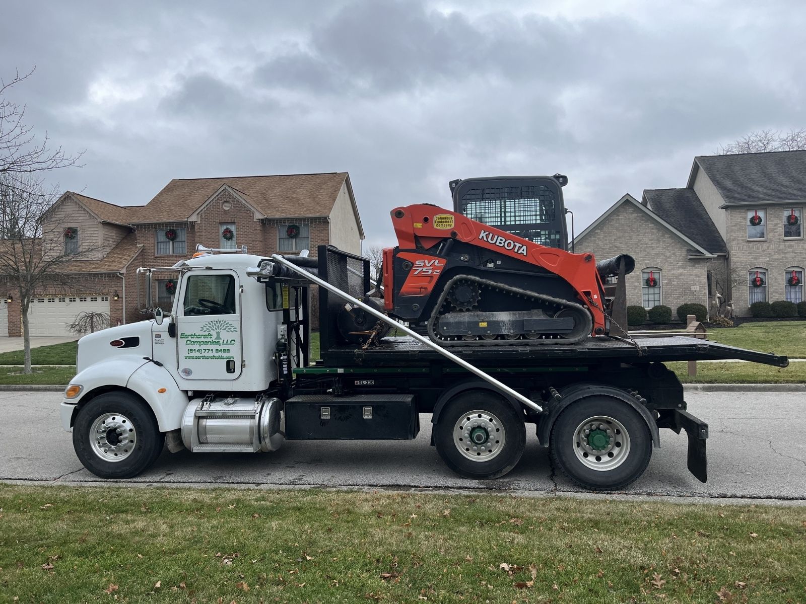 White flatbed truck from Earthworx Companies LLC carrying an orange Kubota SVL75-2 compact track loader parked on suburban street.