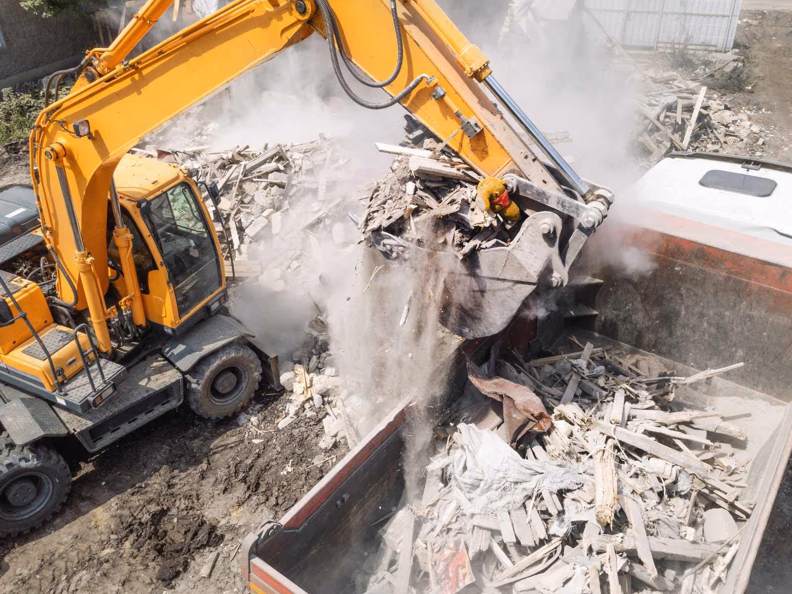 Yellow excavator loading debris including wood and concrete rubble into a large dump truck on a construction site.