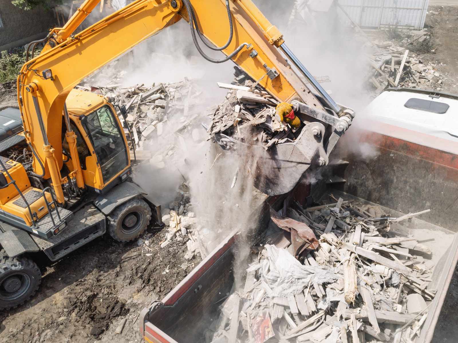Yellow excavator loading debris including wood and concrete rubble into a large dump truck on a construction site.