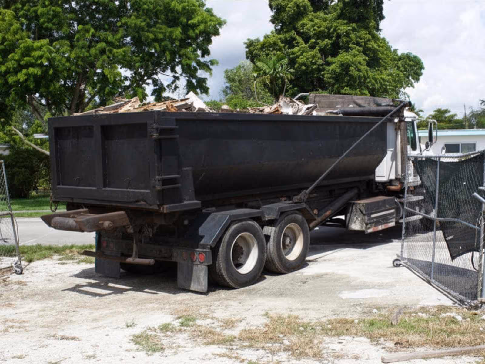 Large black dump truck filled with debris parked on a dirt surface near a chain-link fence and green trees.