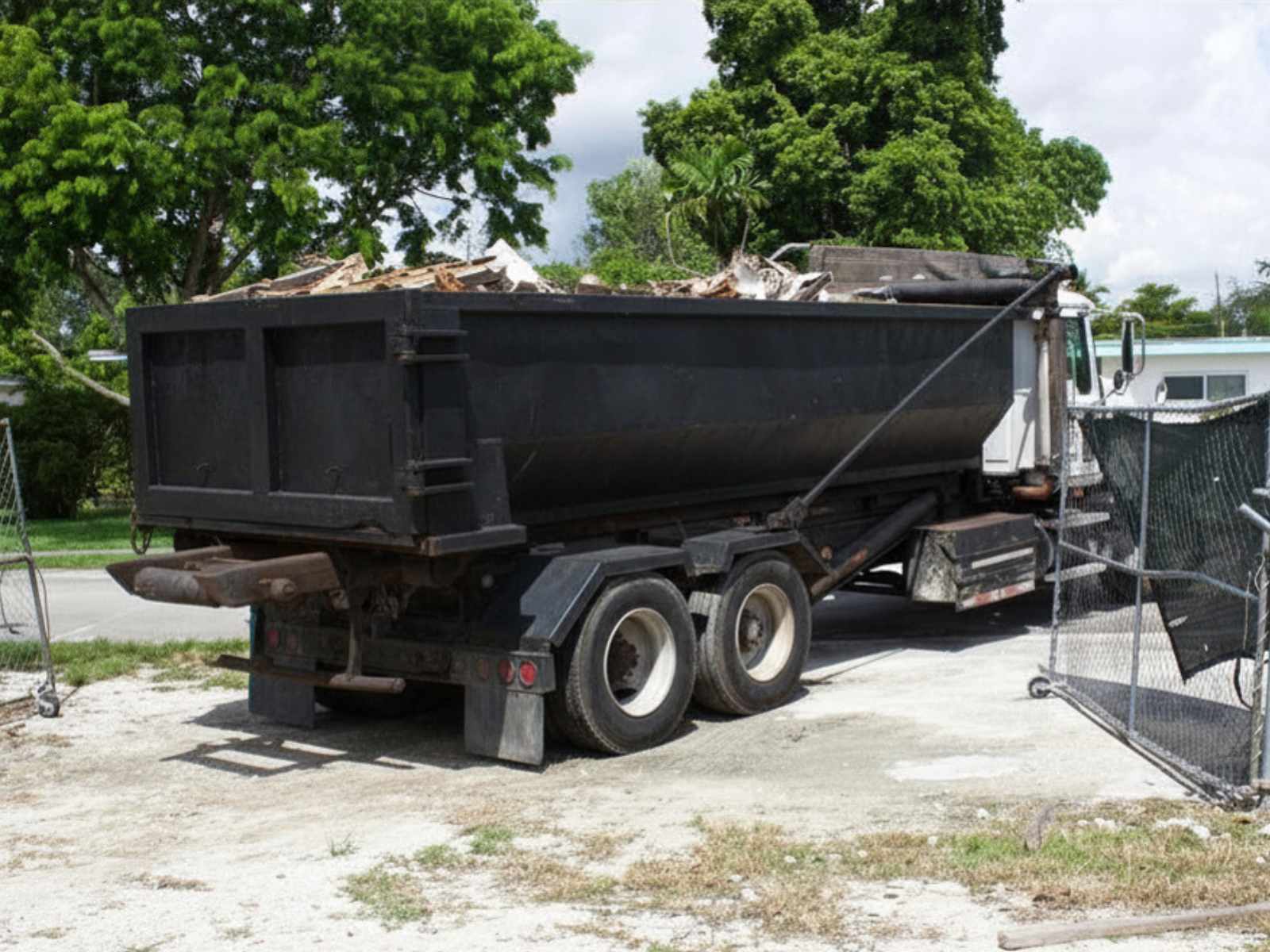 Large black dump truck filled with debris parked on a dirt surface near a chain-link fence and green trees.