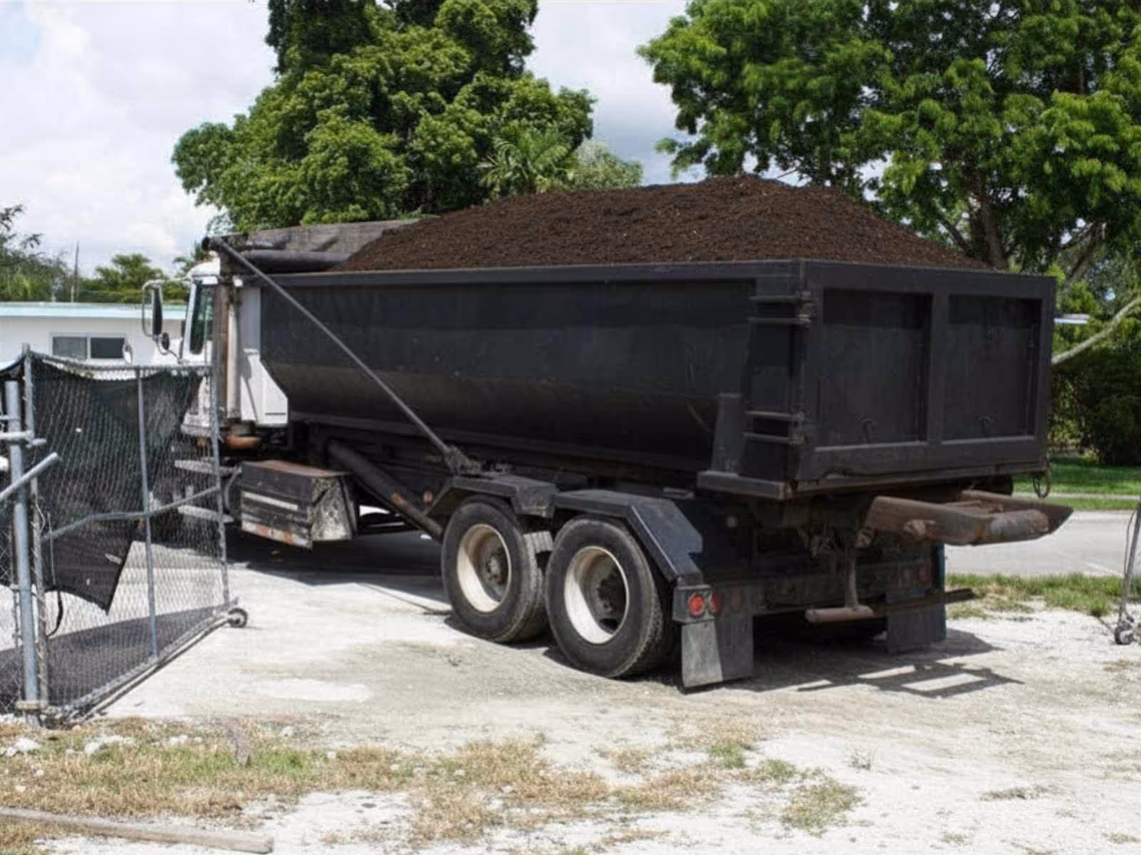 Black dump truck loaded with dark soil parked near a chain-link fence with green trees in the background.