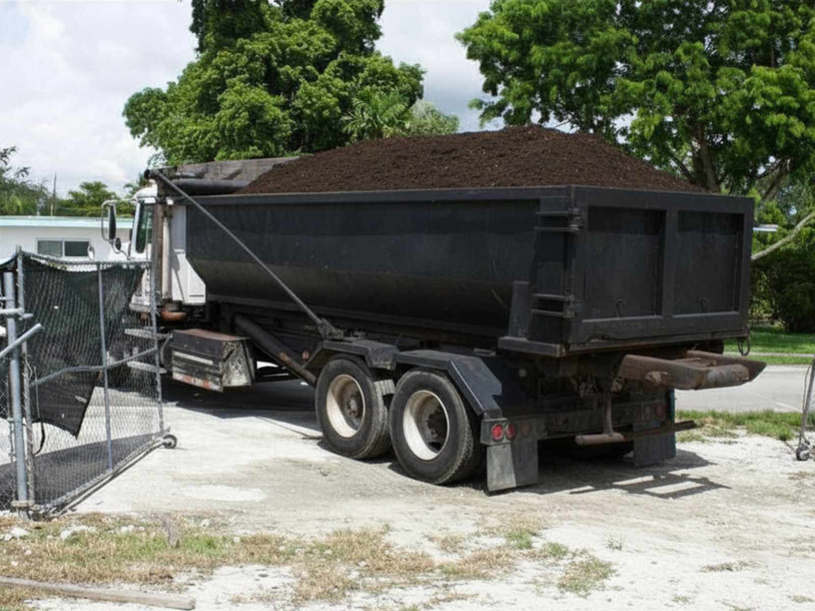 Black dump truck loaded with dark soil parked near a chain-link fence with green trees in the background.