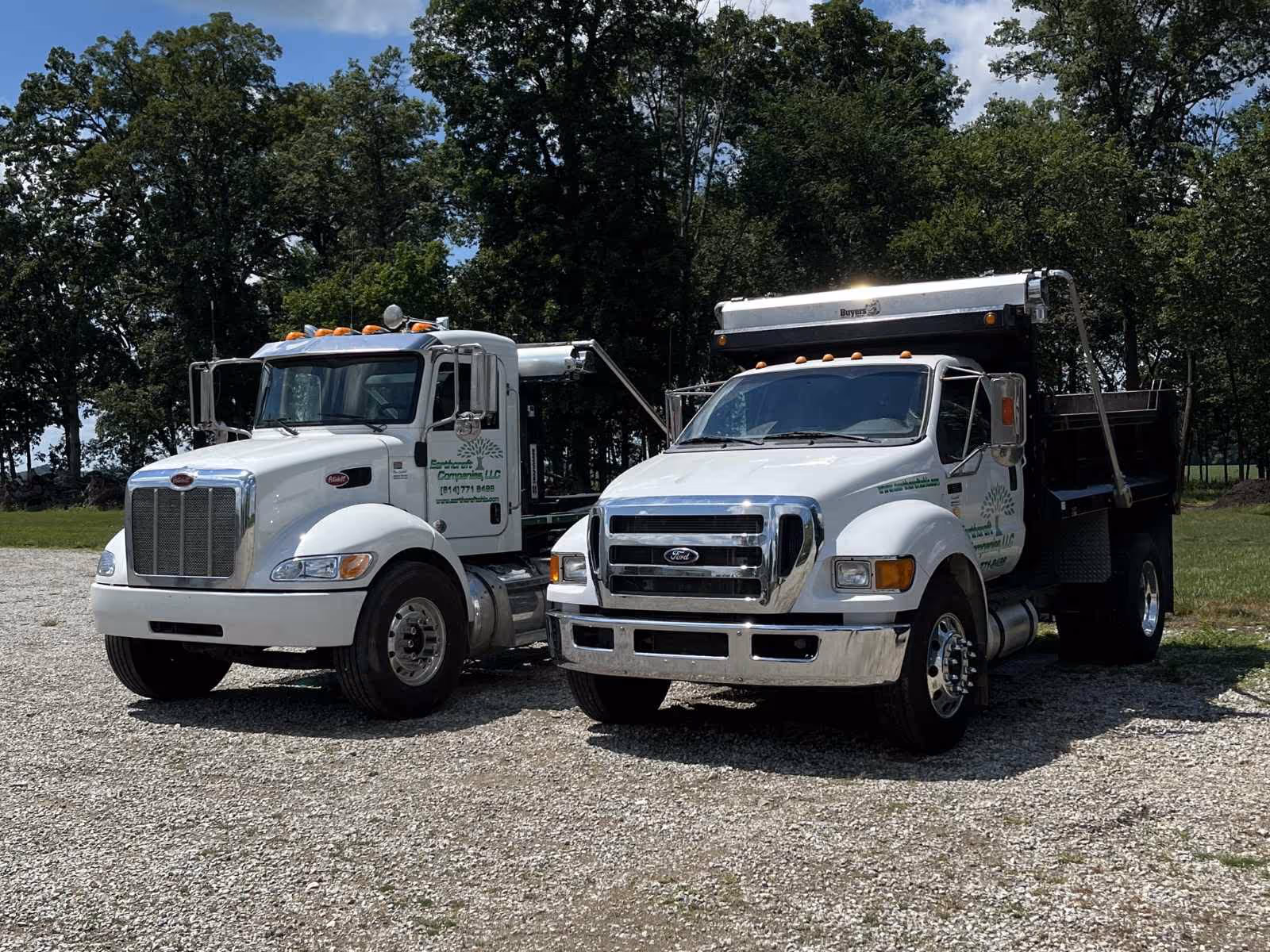 Two white commercial trucks parked on a gravel area with trees and blue sky in the background.