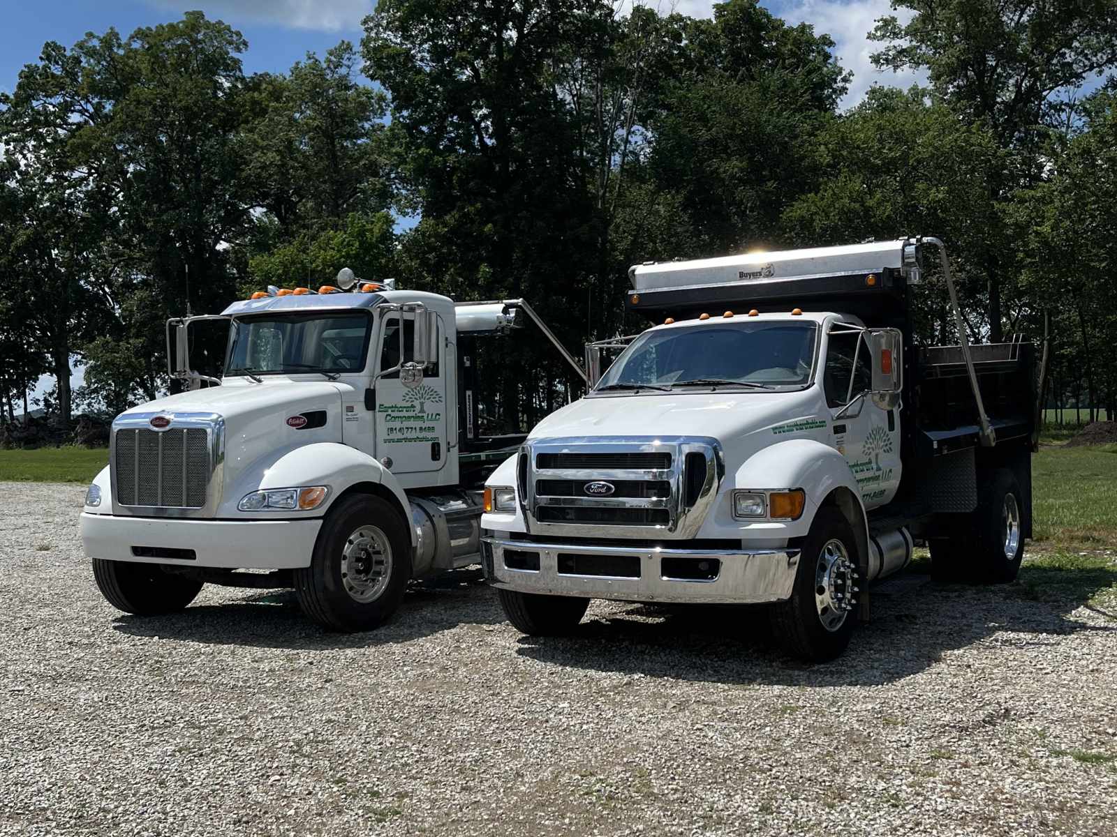 Two white commercial trucks parked on a gravel area with trees and blue sky in the background.