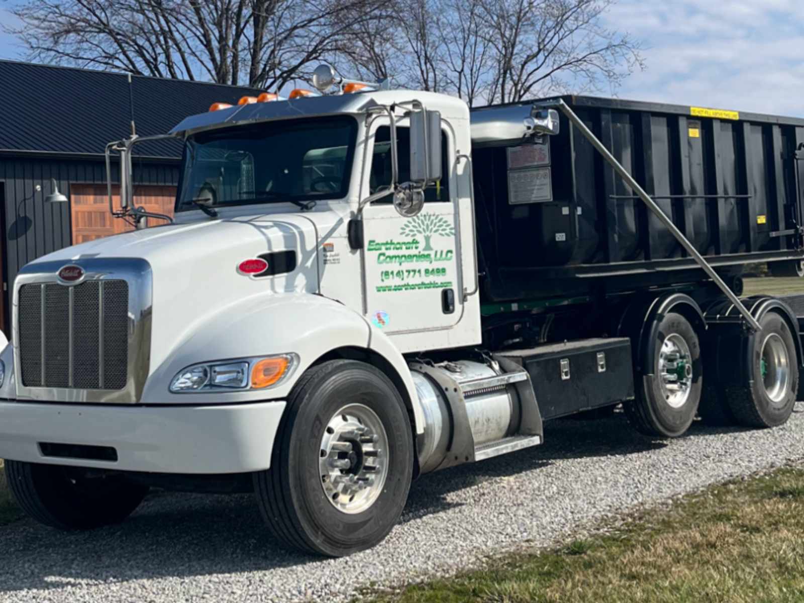 White Peterbilt dump truck with black container and Earthworks Companies LLC logo parked on gravel near a building.