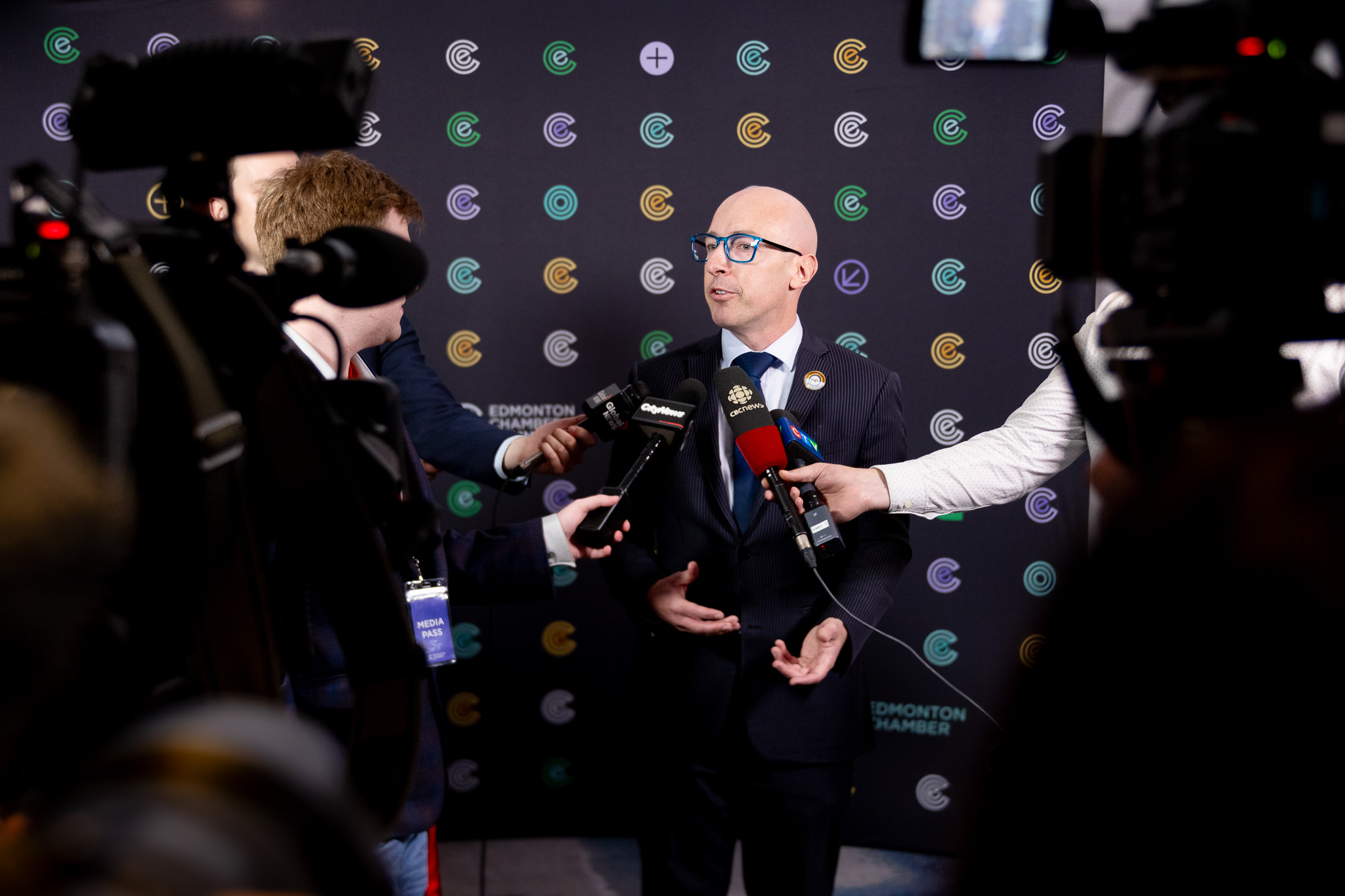 एक सूट लगाएको मान्छे समाचार माध्यमहरूद्वारा गरिएको प्रश्नहरूको जवाफ दिँदै, एडमोन्टन चेम्बर लोगोहरू रहेको पृष्ठभूमिसँग।