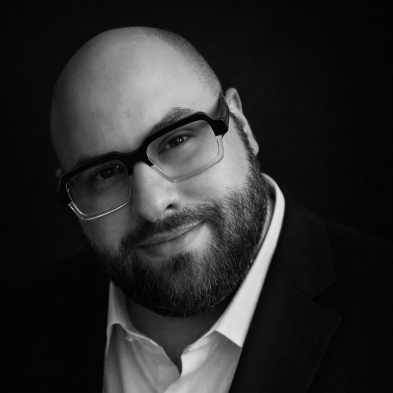 कालो पृष्ठभूमिमा आँखा छोप्ने शैलीका चश्मा लगाएका, दाढी र टाउको नाङ्गो, कालो सूट र सेतो कर्टा लगाएका पुरुषको जवाफ दिने अनुहारको नजिकको तस्वीर।