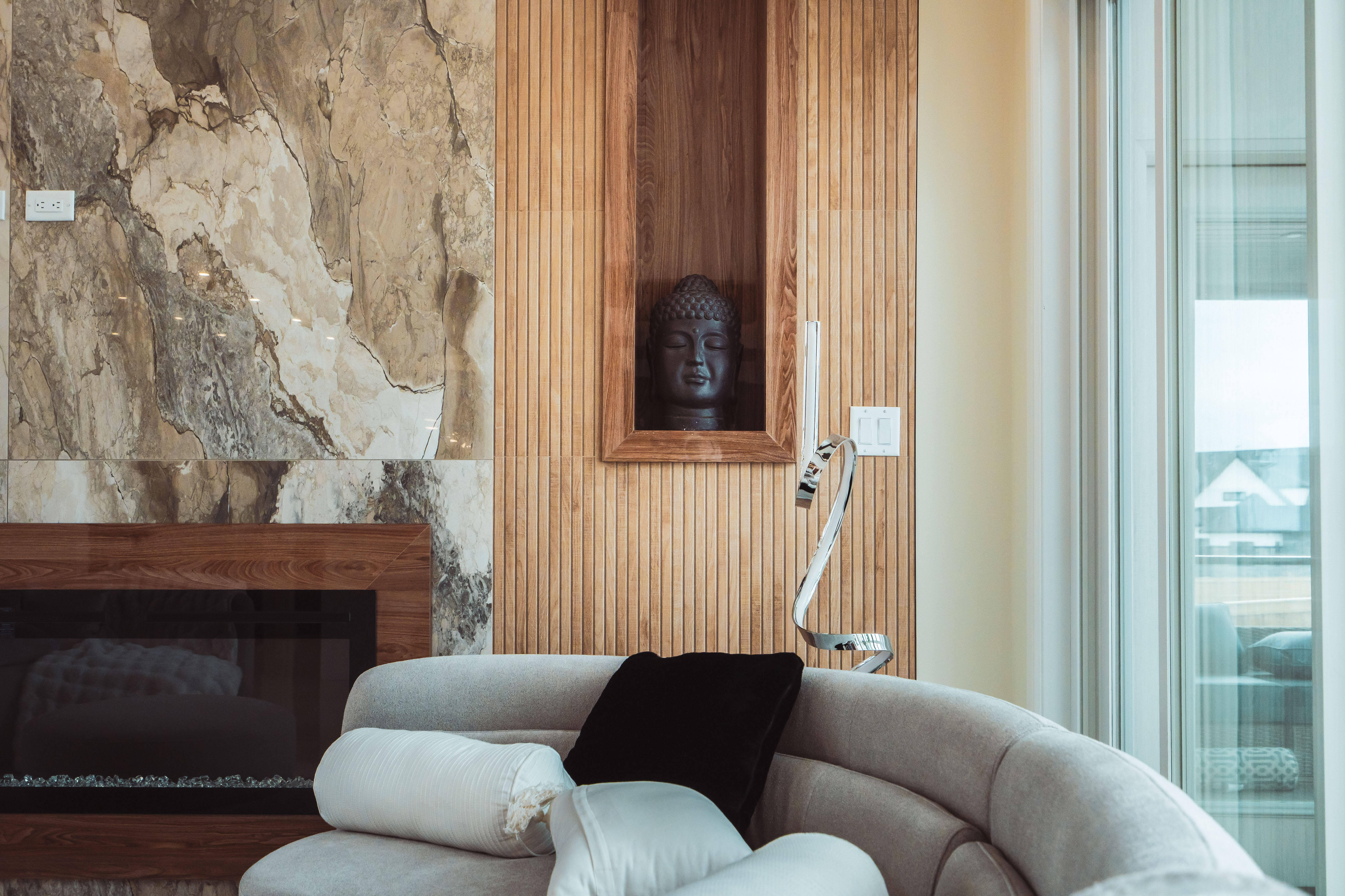 लकडीको प्यानलमा राखिएको बुद्धको कालो प्रस्तर मूर्ति, आँधी मर्मम्य डाडो र हल्का खैरो सोफा सँगको आरामदायक बैठक कोठा।