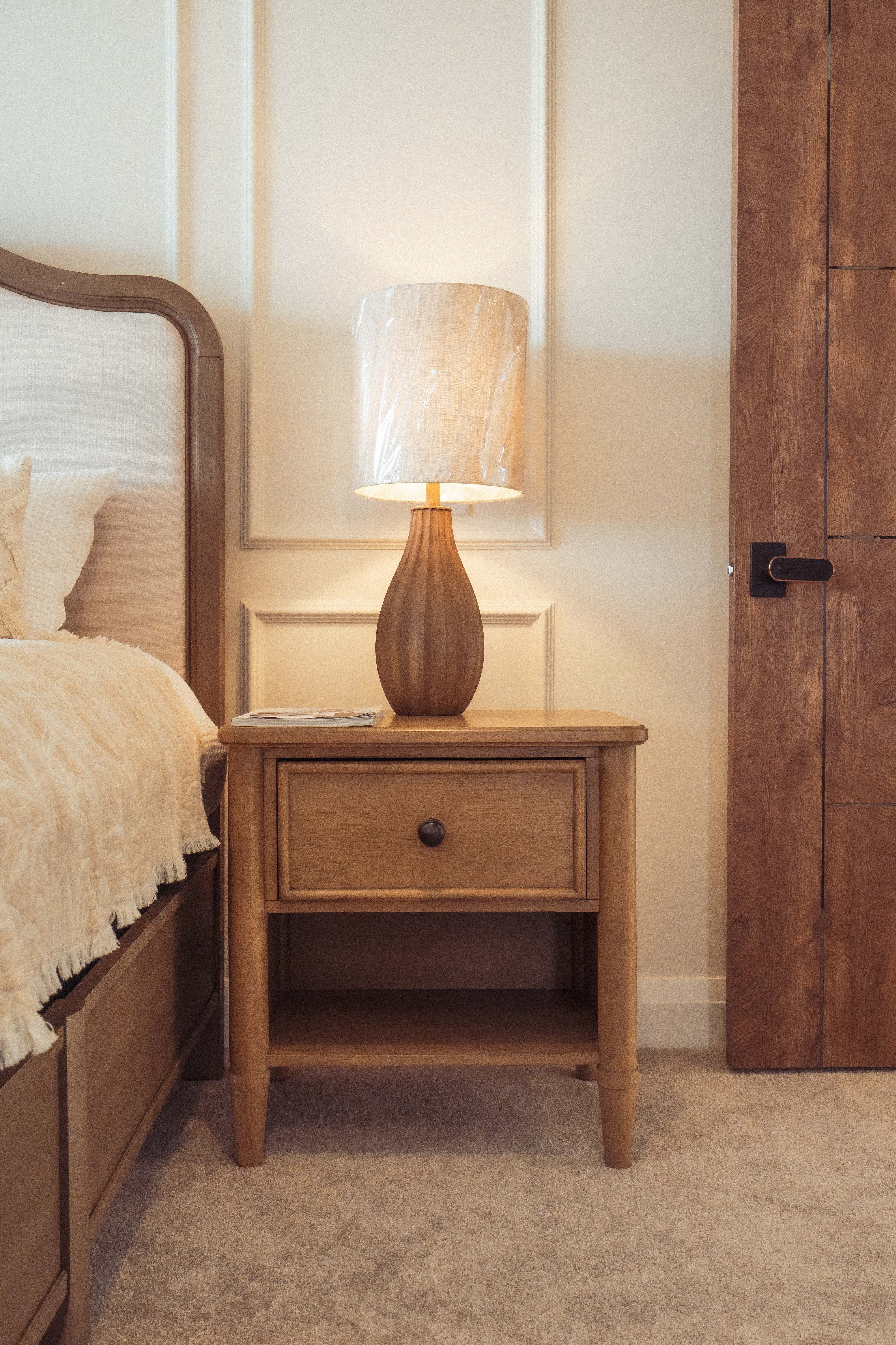 लकडीको झ्यालको छेउमा टेबल ल्याम्प राखिएको नाइटस्ट्यान्ड र आरामदायक सिङ्गो शैयाको भाग।