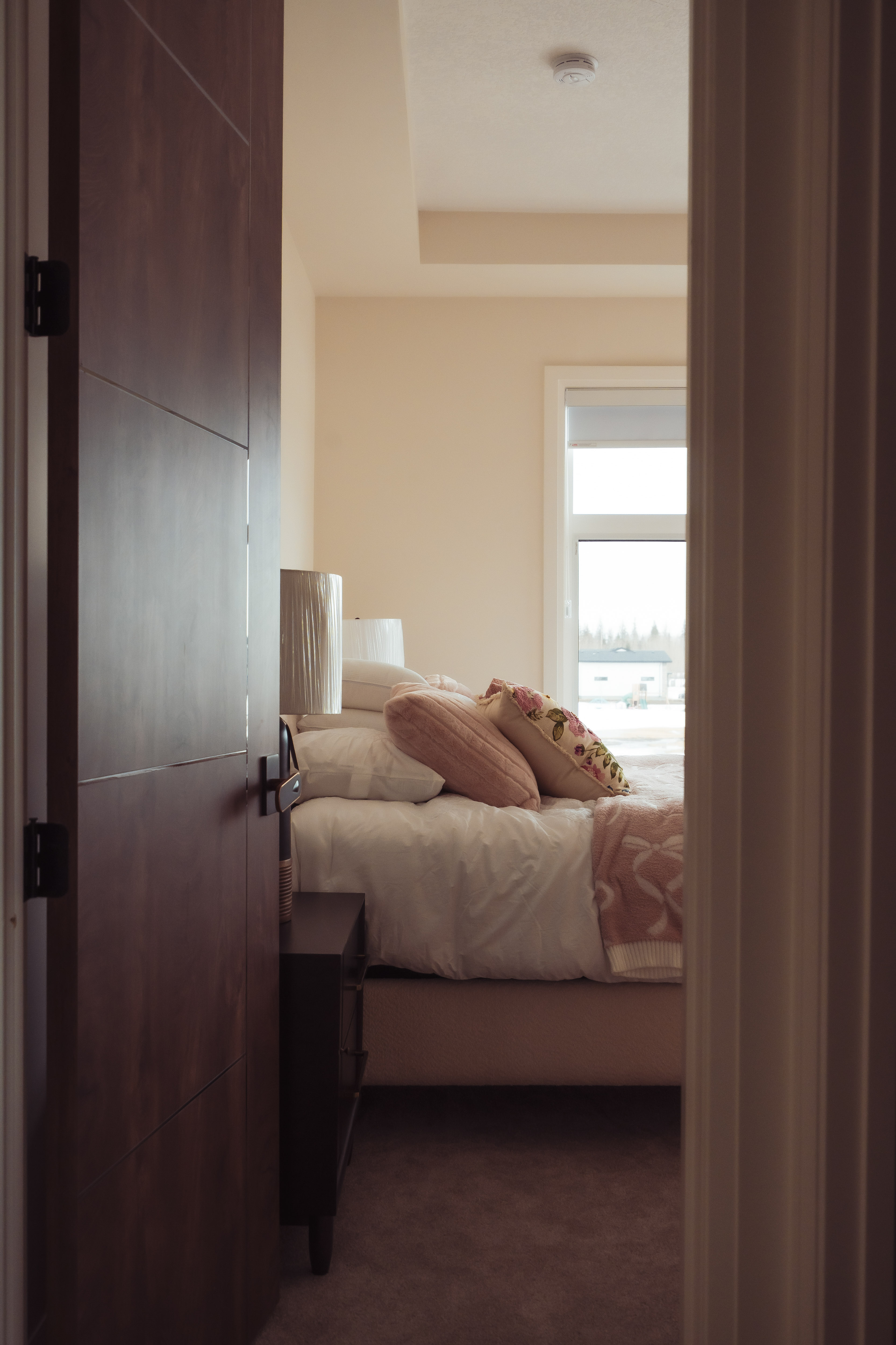 एक सफा बेडरूम जहाँ से सुसज्जित ओछ्यान, गुलाबी रङका तकिया र प्रकाश छरिने झ्याल देखिन्छ।