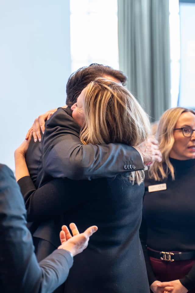 दुई जना व्यक्तिगत रूपमा गाढा आलिंगन गर्दै छन्, पृष्ठभूमिमा अर्की एक महिलाले चश्मा लगाएर दायाँतिर हेर्दै छिन्।