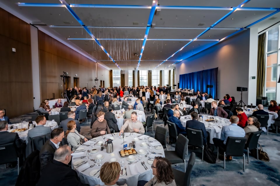 ठूलो सम्मेलन हलमा गोठ्याइएका मेचमा धेरै मानिसहरू भोजन गरिरहेको र कुराकानी गरिरहेको दृश्य।