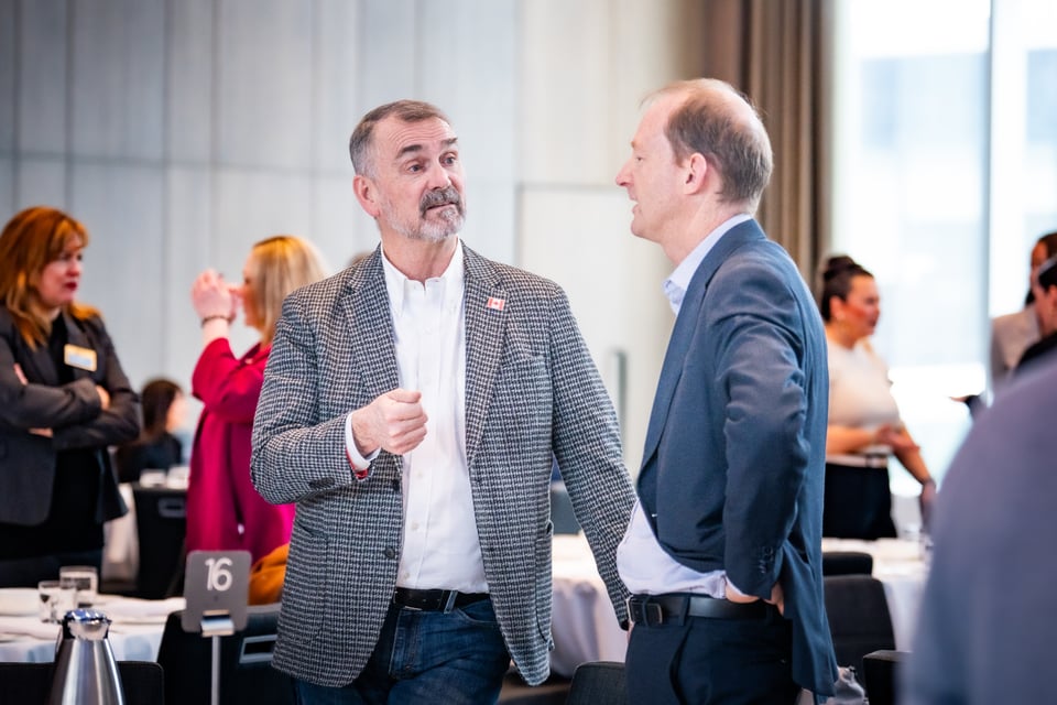 दुई पुरुषहरू भेटघाट गर्दै, एकले गैंर्ड ज्याकेट र सेतो सर्ट लगाएको छ, अर्कोले निलो सूट लगाएको छ, पृष्ठभूमिमा अरू मानिसहरू कुराकानी गरिरहेका छन्।