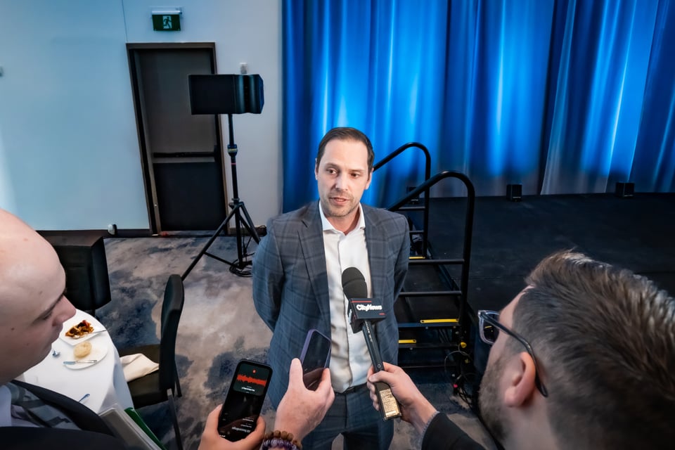 सुट लगाएका एक पुरुष माइक र मोबाइलहरू समातेका पत्रकारहरूसँग कुराकानी गर्दै।