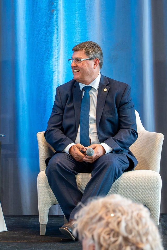 मान्छे नीलो टाई र कालो सूट लगाएर एउटा कुर्सीमा बसेर मुस्कुराउँदै माइक्रोफोन समाउँदै।