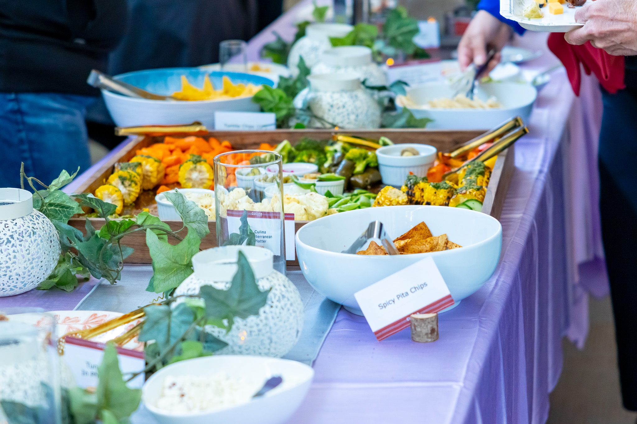 Buffet table with bowls of spicy pita chips, vegetables, and decor on a lavender tablecloth.