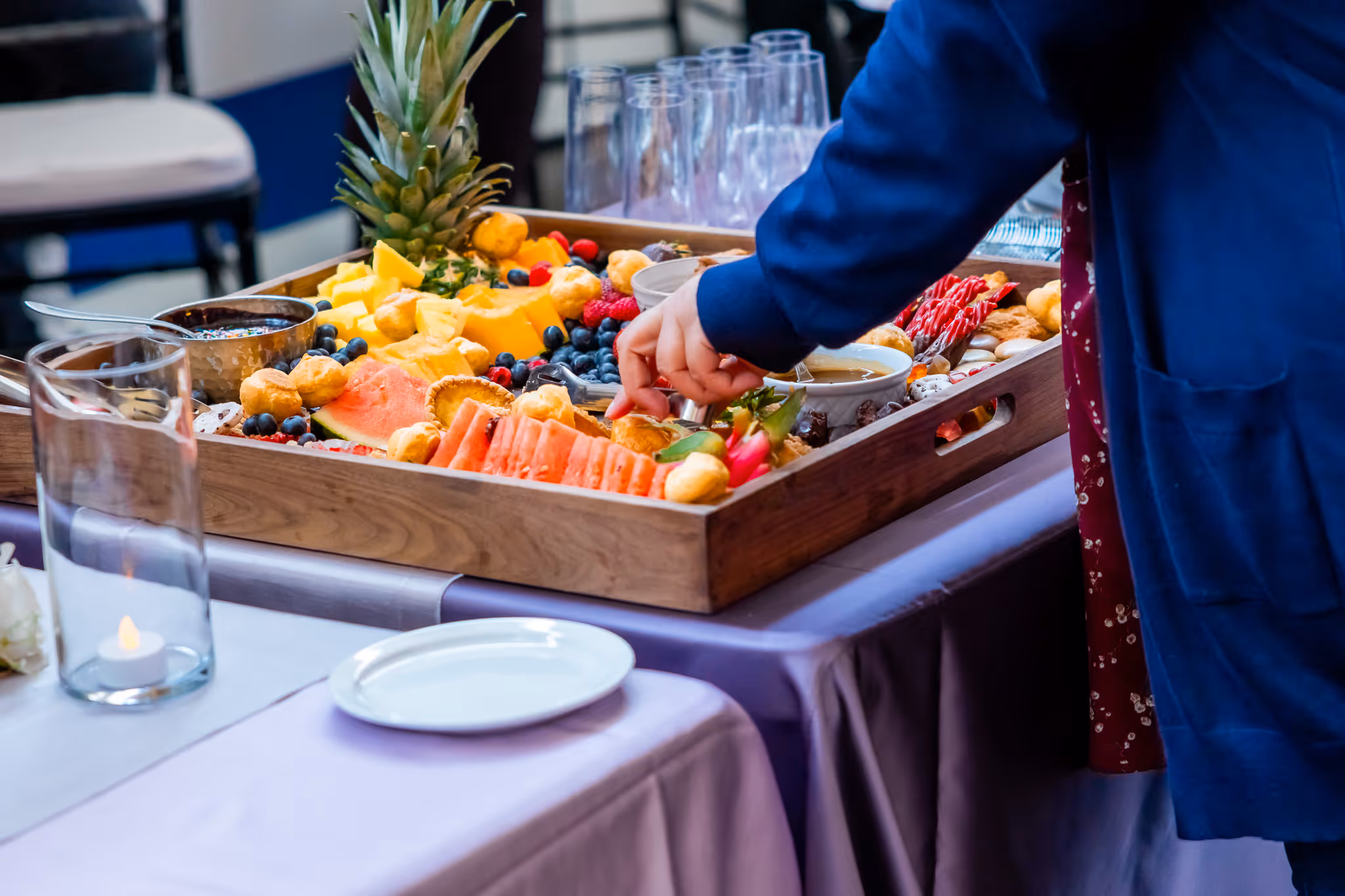 Person in blue jacket reaching for food from a tray with assorted fruits and desserts.
