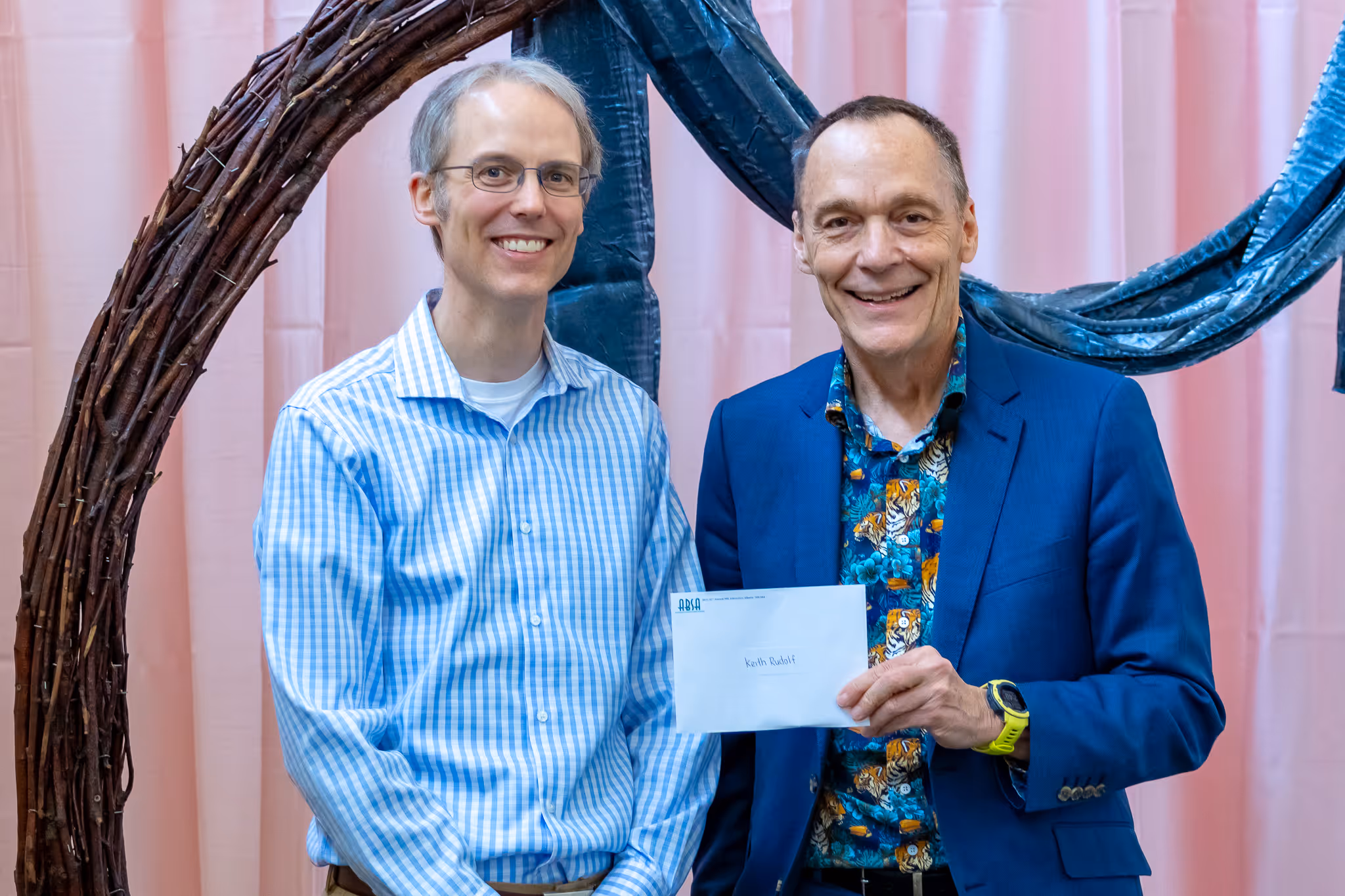 Two men smiling, one holding an envelope labeled Keith Rudolf, standing in front of a pink backdrop.