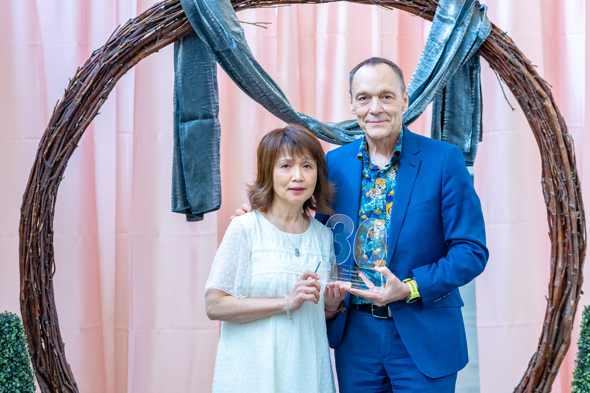 A woman in white dress and man in blue suit hold a 30-year recognition award, standing in front of a wreath.