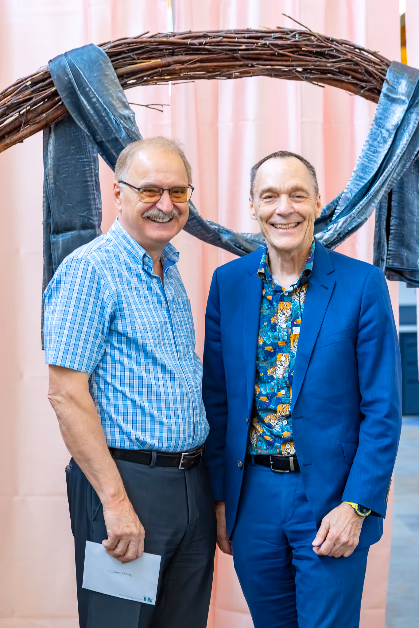 Two smiling men standing indoors with a decorative branch arch wrapped in fabric behind them.