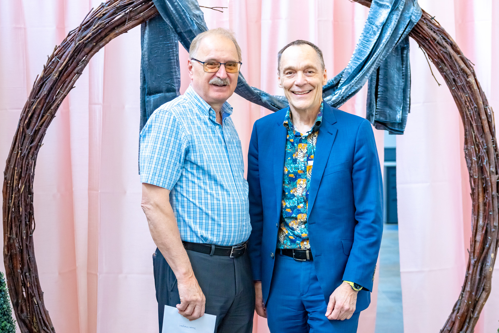 Two middle-aged men smiling, one in a blue checked shirt, the other in a blue suit with a patterned shirt.