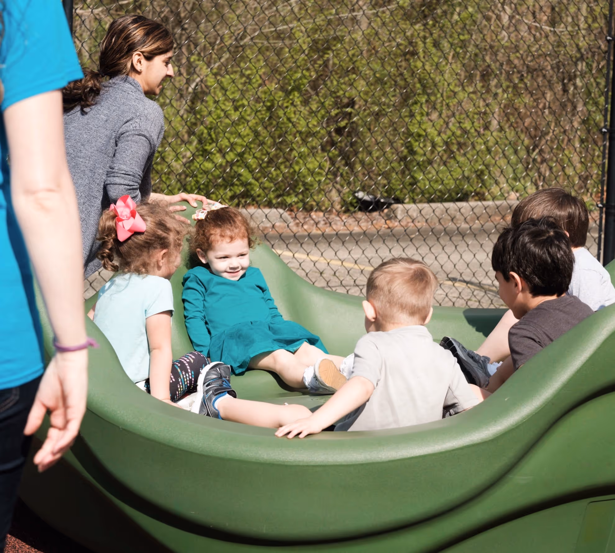 A group of young children sitting together in a large green outdoor play structure with two adults nearby.