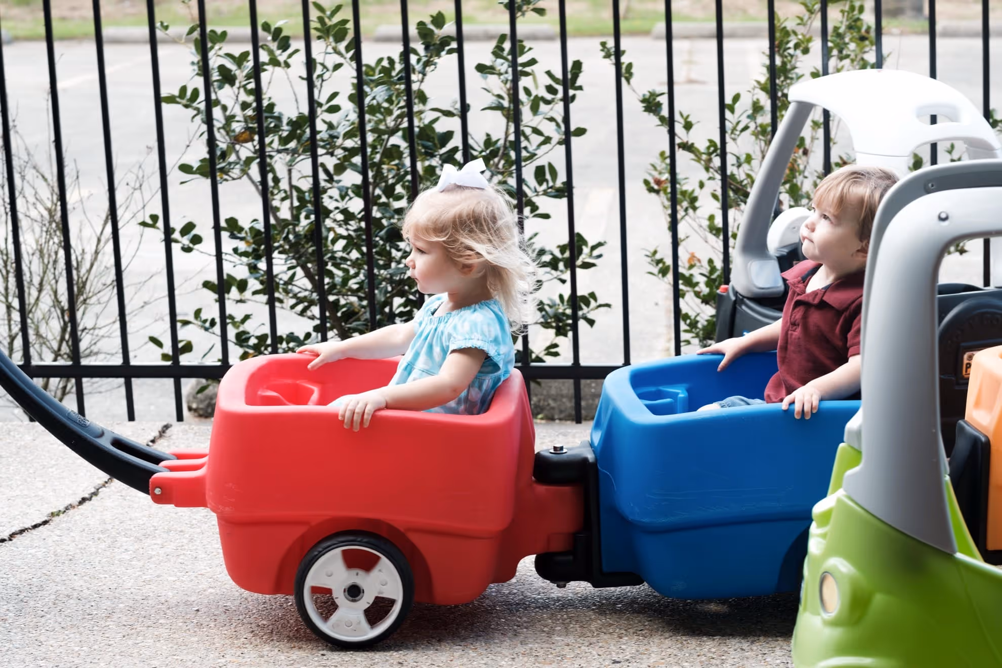 Two toddlers sitting in connected red and blue plastic wagon cars outdoors near a metal fence.
