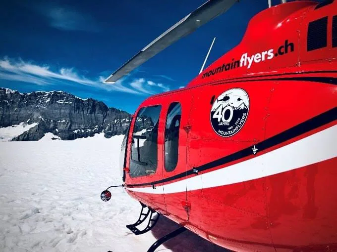 Red helicopter on snow-covered terrain with mountains in the background and a mountainflyers.ch