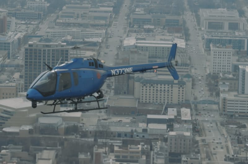 Blue helicopter flying over a city skyline with buildings and streets visible below.