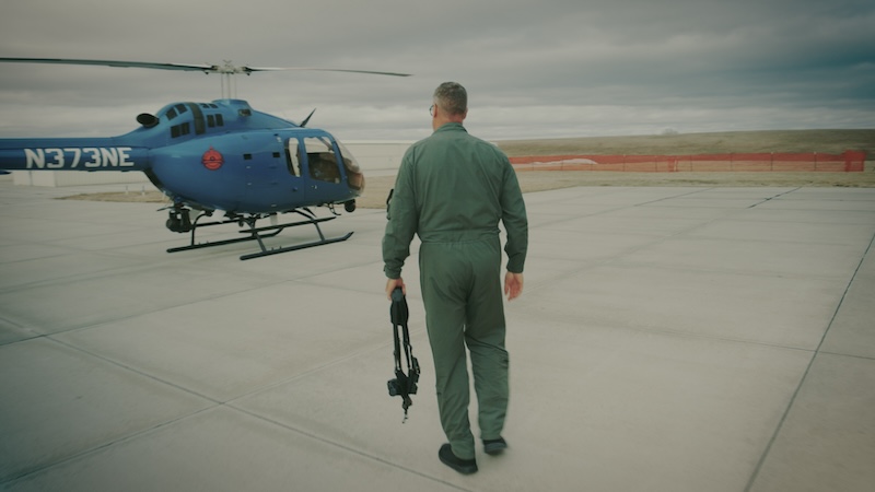 A person in a flight suit walks towards a blue helicopter on an airstrip.