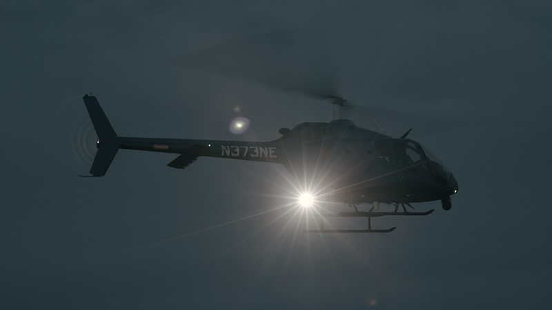 A black helicopter flies in the dark sky, its spotlight illuminating the area below.