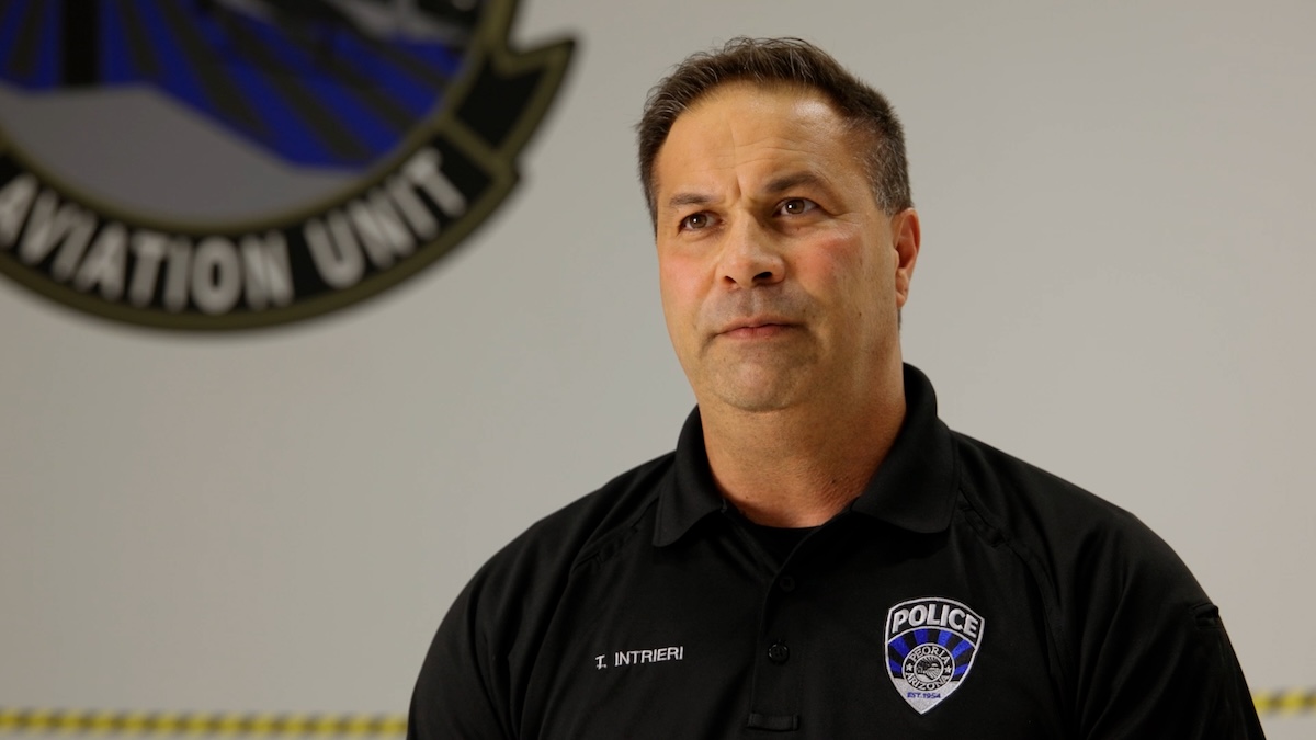 A man in a police uniform speaks in front of a wall featuring an aviation unit emblem.