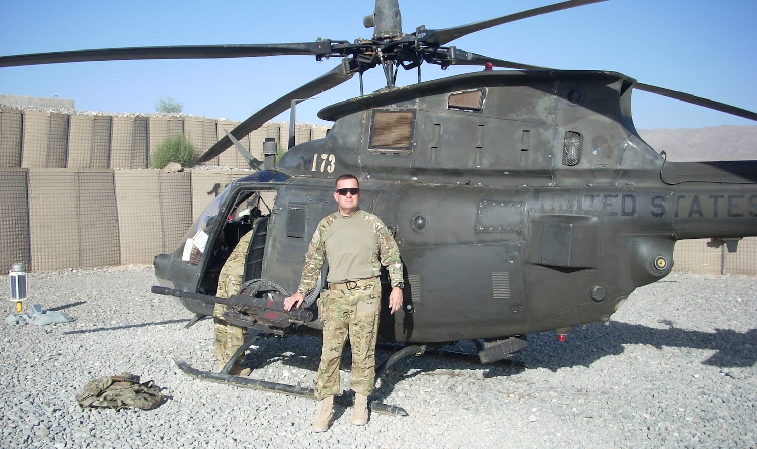 Soldier in military uniform stands beside a black helicopter in a desert setting.