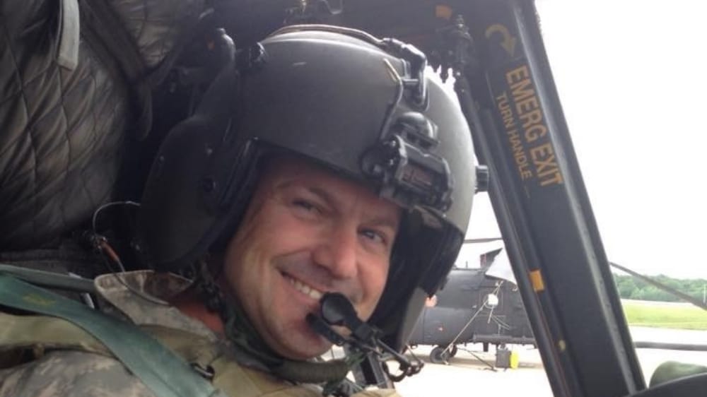 Man wearing a military helmet inside a helicopter, smiling with a headset on. Background shows