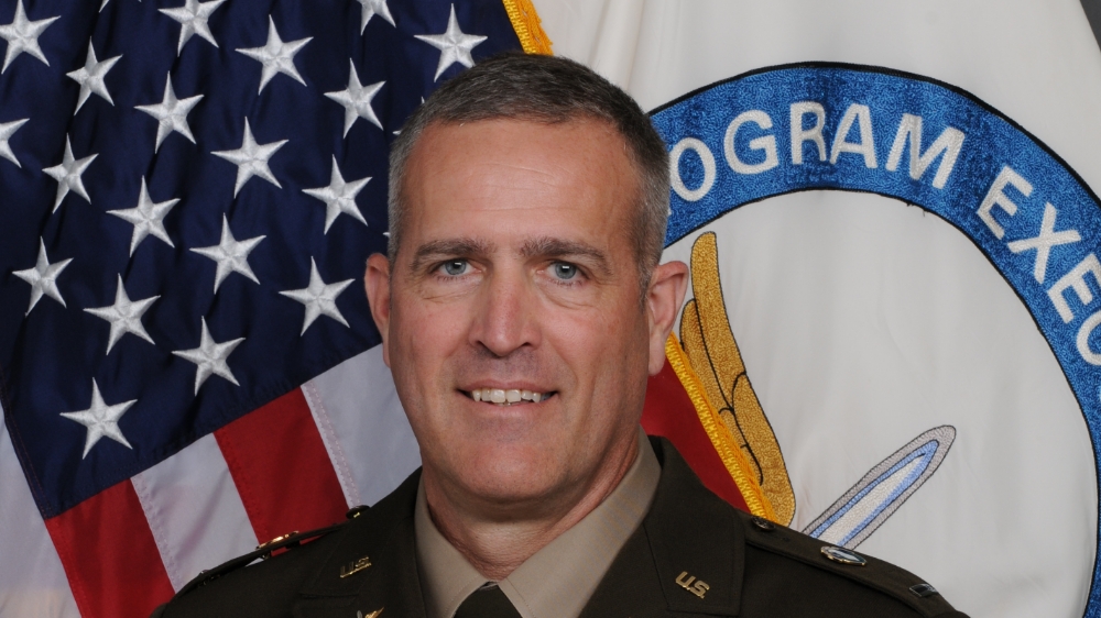 A military officer in uniform smiles in front of the American flag and a program emblem.