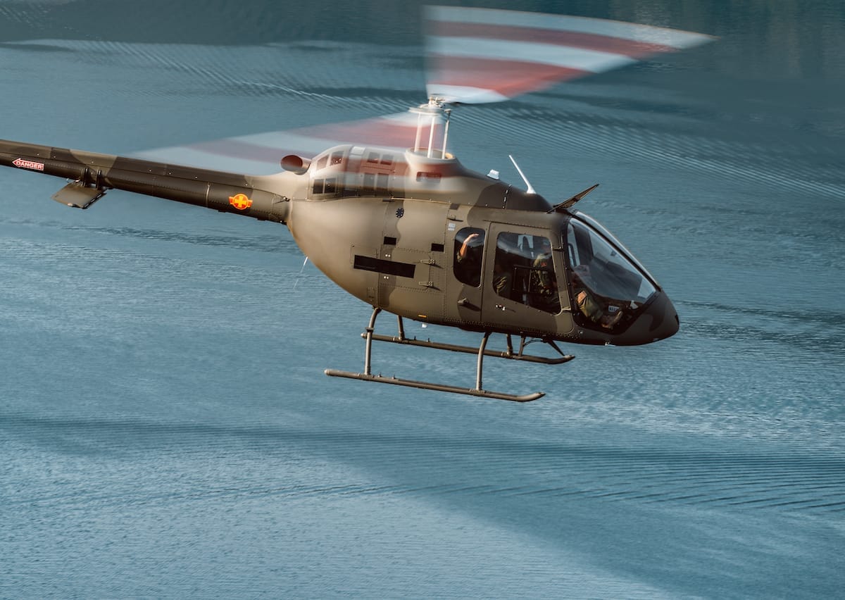 A military trainer helicopter flying over a calm blue body of water. Rotors in motion.