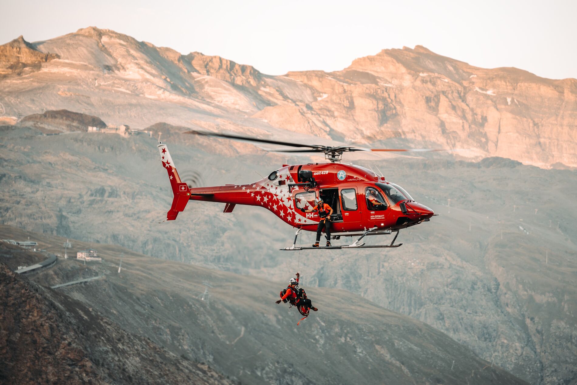 Red rescue Bell 429 airlifting a person with mountains in the background during daylight.