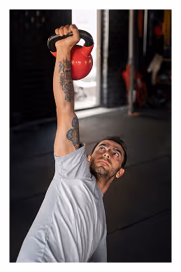 Man with tattoos lifting a red kettlebell overhead while training indoors.