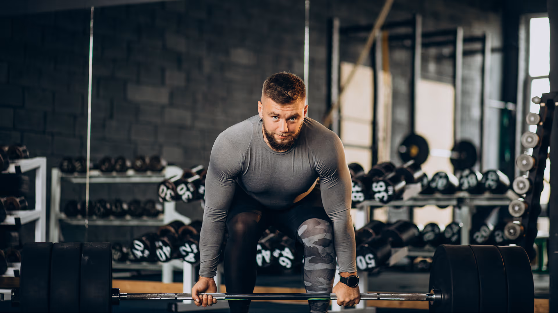 Muscular man in gray long sleeve shirt preparing to lift a heavy barbell in a gym.