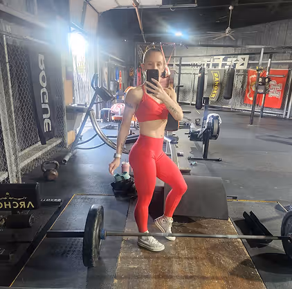 Woman in red workout outfit taking a mirror selfie at a gym with weightlifting equipment around.