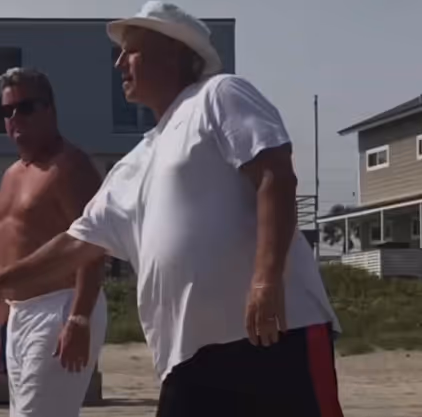 Two men outdoors near beach houses, one wearing a white hat and white shirt.