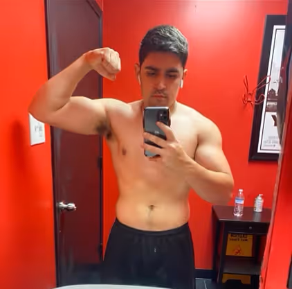 Shirtless young man flexing his arm muscles while taking a mirror selfie in a red-walled room.