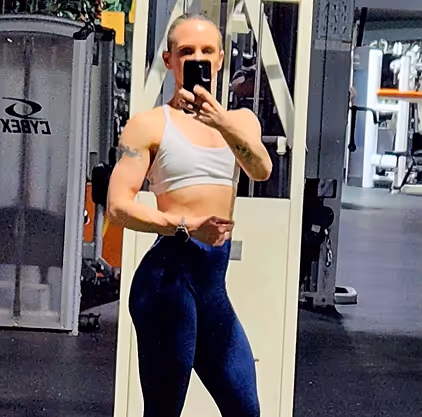 Person in athletic clothing taking a mirror selfie at a gym with exercise equipment in the background.