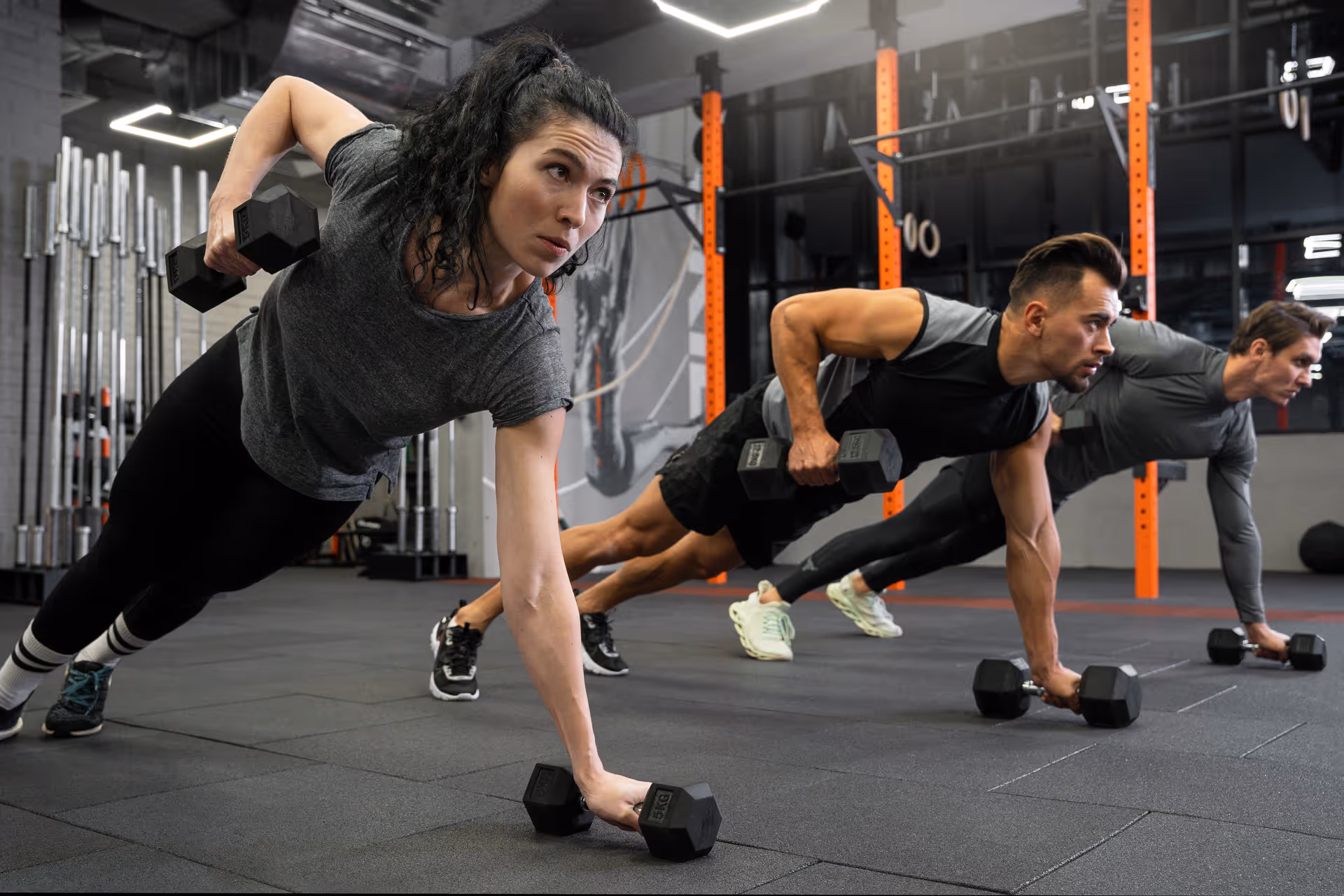 Three people in workout clothes doing dumbbell plank rows in a gym with orange support beams.
